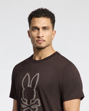 A man with short curly hair wears a black Psycho Bunny Men's Monarc Front Bunny Graphic Tee, made from 100% Pima cotton, featuring stylized bunny ears above crossed bones. He looks directly at the camera against a plain background, showcasing Monarc's unique style.