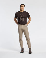 A person stands against a plain background wearing a dark Psycho Bunny B6U211E200 tee made from 100% Pima cotton, light-colored pants, and dark boots.