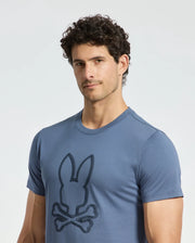 A man with curly hair confidently wears a Psycho Bunny MENS MONARC FRONT BUNNY GRAPHIC TEE - B6U211E200, made of 100% Pima cotton and showcasing a rabbit and crossbones design. He stands against a plain background, looking at the camera.