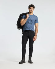 A man stands against a plain backdrop wearing a Psycho Bunny MENS MONARC FRONT BUNNY GRAPHIC TEE in blue, made from 100% Pima cotton. He pairs it with black pants and boots and casually drapes a jacket over his shoulder, highlighting his curly hair.