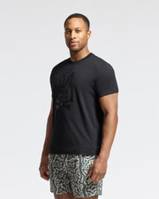 A man confidently wears the Psycho Bunny MENS PAYSON LEATHER PRINT GRAPHIC TEE - B6U213E200, made from soft Pima cotton jersey and featuring a rabbit and skull design. He pairs it with gray patterned shorts against a plain white backdrop, maintaining a neutral expression.