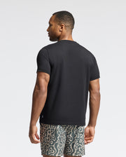 Wearing a Psycho Bunny MENS PAYSON LEATHER PRINT GRAPHIC TEE, the individual is in patterned shorts, showcasing their left side against a plain white backdrop.