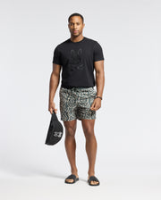 A man in a black Psycho Bunny MENS PAYSON LEATHER PRINT GRAPHIC TEE, patterned shorts, and black slides stands against a plain background, holding a matching black bag.