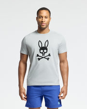 A man wearing a Psycho Bunny graphic tee, the MENS PAYSON LEATHER PRINT GRAPHIC TEE - B6U213E200 in light gray, made from Pima cotton jersey with a black skull and crossbones featuring rabbit ears. He's also wearing blue shorts for a casual look.