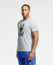 A man in a Psycho Bunny MENS PAYSON LEATHER PRINT GRAPHIC TEE (B6U213E200) with a black skull and crossbones with bunny ears design, paired with blue shorts, stands against a plain background.