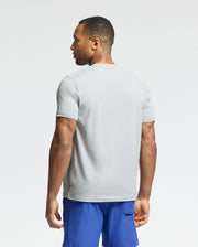 A man in a Psycho Bunny MENS PAYSON LEATHER PRINT GRAPHIC TEE (light gray) and blue shorts stands sideways. The background is plain white.