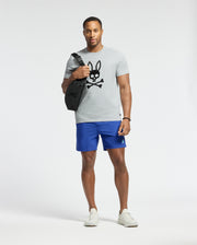 A man confidently stands in athletic attire, wearing a Psycho Bunny MENS PAYSON LEATHER PRINT GRAPHIC TEE in light gray Pima cotton with a bunny and crossbones design, bright blue shorts, white sneakers, and carrying a black shoulder bag, gazing forward against a plain white background.