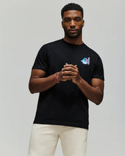 A man stands against a plain background wearing the Psycho Bunny MENS MONTREAL TEE (B6U381F200), a soft black Pima cotton t-shirt with a small colorful chest design, paired with light shorts. He clasps his hands and looks at the camera.