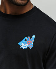 A man wears the Psycho Bunny MENS MONTREAL TEE (B6U381F200), a black Pima cotton shirt with a small chest graphic featuring a pink Psycho Bunny skull and crossbones, blue geometric shapes, and “Mil” stylized text.