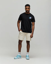 A man with short curly hair and a relaxed, confident posture wears the Psycho Bunny MENS MONTREAL TEE (B6U381F200), styled with beige shorts, white socks, and blue sneakers, embodying Montreal street style against a plain background.