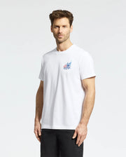 A man with short brown hair and a beard is wearing a Psycho Bunny MENS TORONTO TEE - B6U383F200, featuring a small colorful graphic on the left chest, along with black shorts, standing against a plain light background.