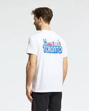 A man with short brown hair faces away, wearing Psycho Bunny’s MENS TORONTO TEE - B6U383F200 in soft Pima cotton, featuring colorful cartoon and city landmark graphics on the back. He pairs it with black shorts.