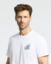 A man with short, wavy brown hair and light stubble wears a plain white Psycho Bunny MENS TORONTO TEE (B6U383F200) featuring a small colorful chest graphic, standing against a plain light gray background.
