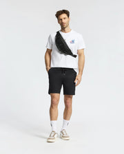 A man with short hair and a light beard stands against a white background, wearing the Psycho Bunny MENS TORONTO TEE - B6U383F200 in soft Pima cotton with bold graphics, black shorts, white socks, beige sneakers, and a black crossbody bag.