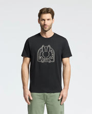 A man wears the Psycho Bunny "MENS MELBOURNE GRAPHIC TEE - B6U410F200," a black Pima Cotton shirt showcasing a white rabbit and bones logo. He pairs it with light green pants featuring an eco-friendly bunny design by the pocket, all on a plain white background.