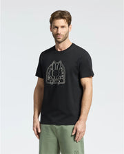 A short-haired, bearded man is wearing the Psycho Bunny MENS MELBOURNE GRAPHIC TEE (B6U410F200) in black, made from eco-friendly Pima Cotton. It features a white "Psycho Bunny" graphic and pairs it with light green pants showing a small bunny logo on a plain white background.