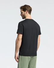 A man with short brown hair stands on a white background, looking right, wearing a black Psycho Bunny MENS MELBOURNE GRAPHIC TEE (B6U410F200) featuring a subtle bunny graphic, paired with green pants made from eco-friendly Pima cotton.