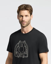 A man with short brown hair and a beard wears the edgy Psycho Bunny MENS MELBOURNE GRAPHIC TEE, B6U410F200. The black Pima Cotton shirt features a stylized bunny graphic with crossbones and "Psycho Bunny" text, standing out against a plain light gray background.