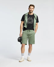 A man in a black Psycho Bunny MENS MELBOURNE GRAPHIC TEE, paired with green shorts and a green sweatshirt draped over his shoulders. He holds a black cap, completing the look with white sneakers and striped socks.