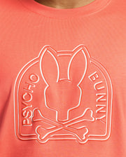 The Melbourne tee by Psycho Bunny, made from eco-friendly Pima Cotton in coral, features an embossed bunny head with crossbones design and "Psycho Bunny" printed on the sides.