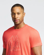 A person in a coral pink Psycho Bunny MENS MELBOURNE GRAPHIC TEE (B6U410F200) made of eco-friendly Pima Cotton, featuring a raised bunny and crossed bones design. They have short hair and stand against a plain, light background.