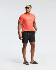 A man in a Psycho Bunny MENS MELBOURNE GRAPHIC TEE - B6U410F200, black shorts, black slides, and sunglasses strikes a relaxed pose against a plain background.