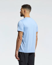 A man with short curly hair stands facing away from the camera, wearing the Psycho Bunny MENS MELBOURNE GRAPHIC TEE - B6U410F200 in light blue and black pants against a plain light background.
