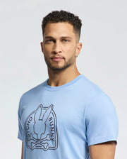 Someone wearing a Psycho Bunny MENS MELBOURNE GRAPHIC TEE (B6U410F200) in light blue stands against a plain background. Made from eco-friendly Peruvian Pima cotton, the tee features a stylized bunny and crossbones design in dark print.