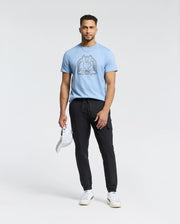 A man confidently stands against a plain background, wearing a Psycho Bunny Peruvian Pima Melbourne tee in light blue with a graphic design, paired with black joggers and white sneakers. He holds an eco-friendly white cap in his left hand.