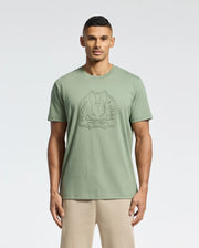 A man stands facing forward in a light green Psycho Bunny MENS MELBOURNE GRAPHIC TEE (B6U410F200) with a large embossed bunny and crossbones logo, paired with beige pants, against a plain light background.