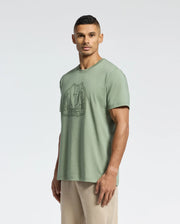 A man in a sage green Psycho Bunny MENS MELBOURNE GRAPHIC TEE (B6U410F200), made from eco-friendly Peruvian Pima cotton, and light pants stands against a plain light background, glancing slightly to the side.