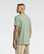 A man with short dark hair stands facing away, wearing the Psycho Bunny MENS MELBOURNE GRAPHIC TEE - B6U410F200 in light green and beige pants against a plain, light background.