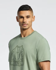 A man with short dark hair wears the Psycho Bunny MENS MELBOURNE GRAPHIC TEE (B6U410F200), a light green, eco-friendly shirt featuring a raised "Psycho Bunny" logo. He stands against a plain light background, looking slightly aside.