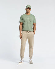A man faces forward in a sage green Psycho Bunny MENS MELBOURNE GRAPHIC TEE - B6U410F200, featuring Bugs Bunny and made from eco-friendly Peruvian Pima cotton, paired with beige jogger pants and white sneakers against a plain light background.