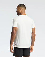 A person wearing a Psycho Bunny MEN'S NEW CASTLE EMBROIDERED GRAPHIC TEE, made from Pima cotton, and black shorts is standing and facing away from the camera against a light gray background.