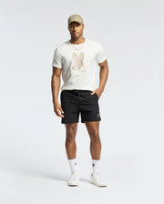 A person in a beige cap and a Psycho Bunny MENS NEW CASTLE EMBROIDERED GRAPHIC TEE featuring a bunny design poses against a neutral backdrop. They wear black shorts, white socks, and sneakers with their hands resting casually by their sides.