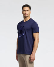 A man in a navy blue Psycho Bunny MENS NEW CASTLE EMBROIDERED GRAPHIC TEE (B6U415F200) with a bunny and crossbones logo stands against a light gray background. He has short, curly hair and wears beige pants. The tee is made from soft Pima cotton for comfort and style.