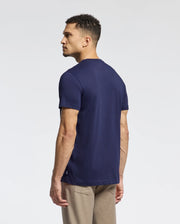 A short, curly-haired person stands leftward against a white backdrop, wearing beige pants and a Psycho Bunny navy graphic tee named "MENS NEW CASTLE EMBROIDERED GRAPHIC TEE - B6U415F200.