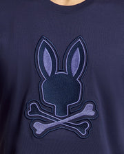The Psycho Bunny MENS NEW CASTLE EMBROIDERED GRAPHIC TEE in navy blue features a soft Pima cotton fabric with an embroidered bunny head and crossed bones, using lighter blue shades for contrast.
