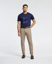 A man stands against a plain background, wearing the Psycho Bunny MENS NEW CASTLE EMBROIDERED GRAPHIC TEE in navy blue and khaki pants. He completes his look with brown shoes and a belt, maintaining a casual posture and neutral expression.