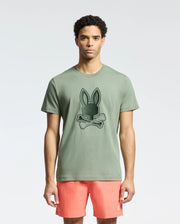 Sporting Psycho Bunny's MENS NEW CASTLE EMBROIDERED GRAPHIC TEE, a green Pima cotton top with a dark bunny and crossbones graphic, he pairs it with coral shorts. His curly hair contrasts the plain background, creating a casual yet striking look.