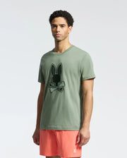 A man with curly hair wears a Psycho Bunny green Pima cotton T-shirt (MENS NEW CASTLE EMBROIDERED GRAPHIC TEE - B6U415F200) featuring a black bunny and crossbones. He pairs it with pink shorts, with two-tone embroidery enhancing the tee against a plain white background.