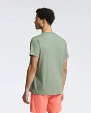 A man with short, curly hair wears a light green Psycho Bunny MENS NEW CASTLE EMBROIDERED GRAPHIC TEE and salmon-colored shorts against a plain white background, highlighting the two-tone embroidery along the shirt's hem.