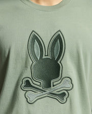 The MENS NEW CASTLE EMBROIDERED GRAPHIC TEE by Psycho Bunny features a playful twist on the classic skull and crossbones symbol with a large dark green stylized bunny head graphic above crossed bones, set on a soft light green Pima cotton fabric.