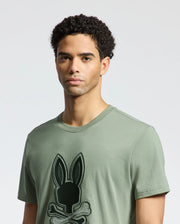 A man wearing the Psycho Bunny MENS NEW CASTLE EMBROIDERED GRAPHIC TEE - B6U415F200, featuring a black bunny and crossbones graphic, stands against a plain white background.
