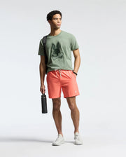 A young man sports a Psycho Bunny MENS NEW CASTLE EMBROIDERED GRAPHIC TEE in green, paired with coral drawstring shorts and white sneakers. He holds a water bottle and has a strap over his shoulder against a plain gray background.