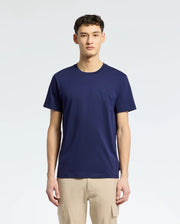 A person wearing a Psycho Bunny MENS NICHOLSON PB CHEST GRAPHIC TEE - B6U416F200 in navy blue, featuring an embroidered logo, pairs it with beige pants against a light gray background. Made from soft Pima cotton, this tee blends comfort with understated style.