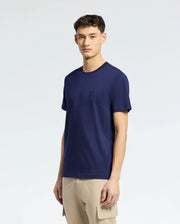 A person in a navy blue Psycho Bunny MENS NICHOLSON PB CHEST GRAPHIC TEE, featuring an embroidered logo on the upper left, pairs it with beige cargo pants while standing against a plain white background.