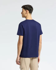A person stands facing away from the camera, wearing a Psycho Bunny MENS NICHOLSON PB CHEST GRAPHIC TEE (B6U416F200) in navy blue and beige cargo pants, highlighting an embroidered logo on the tee against a light-colored background.