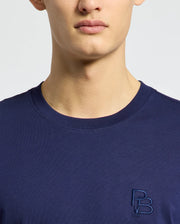A person is wearing a dark blue Psycho Bunny MENS NICHOLSON PB CHEST GRAPHIC TEE, featuring an embroidered "PB" logo on the chest. The background is plain and light, and the image is cropped from the shoulders up.